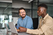© insta_photos - Two busy happy diverse male employees using laptops computers talking in office. Professional young business men workers team having conversation working on project sharing ideas sitting at work desk.