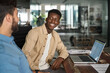 © insta_photos - Happy African male employee having conversation with Latin colleague at work. Smiling friendly diverse workers team talking working together using laptop computer sharing ideas at workplace in office.