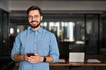 Happy young Latin business man holding phone standing at work in office. Smiling professional businessman company executive, employee or business owner looking at camera with smartphone. Portrait.