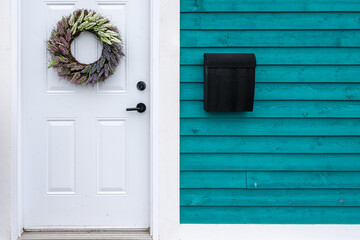 A teal-colored exterior wall of a house has a white door with a silver flower wreath hanging on it. A black metal mailbox and a large four-pane double-hung window are also on the colorful wall.