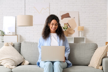 Wall Mural - Pretty young African-American woman with laptop sitting on sofa at home