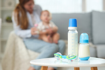 Canvas Print - Bottle of milk and rattle on table against mother with baby at home, closeup