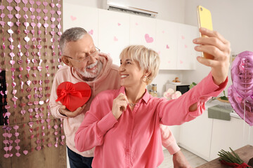 Wall Mural - Happy mature couple with gift box and mobile taking selfie in kitchen at home. Valentine's Day celebration