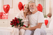 © Pixel-Shot - Happy mature couple with bouquet of roses sitting on bed at home. Valentine's Day celebration