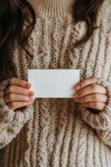 A person wearing a cozy, knitted sweater holds a blank business card mockup in their hands. The focus is on the card and the hands, with a soft, warm background.