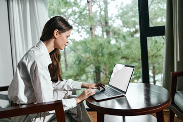 Canvas Print - Young woman working on a laptop in a bright, modern room, dressed in a casual white outfit, with natural light and greenery outside, promoting productivity and focus