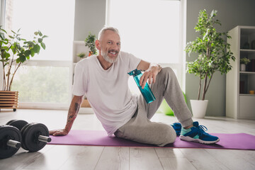 Wall Mural - Smiling senior man in activewear relaxing on a yoga mat indoors with dumbbells and plants around him