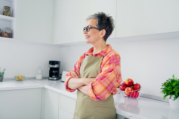 Wall Mural - Elderly woman in kitchen wearing apron and plaid shirt enjoys leisure time at home with fresh apples and greenery around her