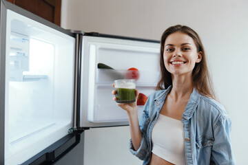 Canvas Print - Young woman smiling while holding a green smoothie in front of an open refrigerator filled with fresh vegetables, representing healthy eating and lifestyle choices