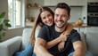 © natakot - A happy couple sits together on a soft sofa, radiating joy and affection in a bright, inviting living room. The woman, with long brown hair, hugs the man from behind, both smiling broadly, showcasing