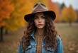 © Vibudhaart - A woman with long curly hair and a brown hat stands in front of a background of fall foliage.