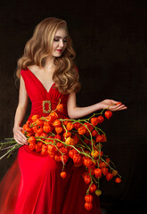 Beautiful young girl - blonde with long light red hair in long red dress holds big bouquet of dry orange flowers of physalis on black background. Studio photography. Autumn art portrait.