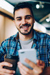 © BullRun - Portrait of cheerful male blogger using telephone for chatting with followers and confirming new friends enjoying social media, smiling student looking at camera holding smartphone on coffee break
