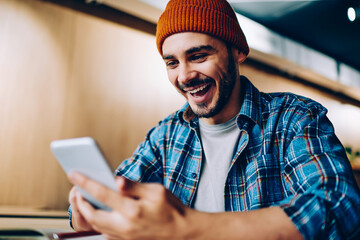 Overjoyed male in hat surprised with victory in online competition in social networks getting notification on cellular, amazed hipster guy happy about winning discount for online courses studying