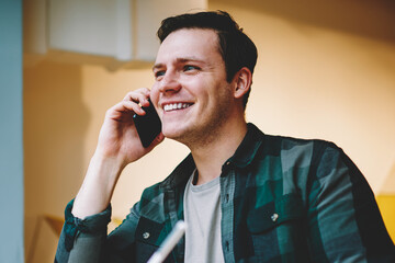 Cheerful hipster guy writing in notebook and talking on telephone sitting at coworking space, happy young man making notes of information during consultancy about credit satisfied with banking service