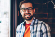 © BullRun - Portrait of successful bearded hipster blogger in eyeglasses smiling at camera while updating profile on smartphone via 4G internet.Happy young man in casual stylish shirt holding mobile phone in hand