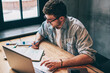 © BullRun - Hipster guy pondering on idea for essay sitting at wooden table with laptop device and looking on textbook, smart male freelancer spending time for distance job thinking about business indoors