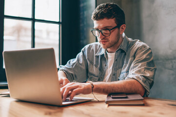 Concentrated male it professional typing program code via application on laptop computer, young serious hipster guy in eyewear searching information using internet, business and freelance concept