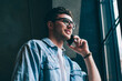 © BullRun - Positive male user in spectacles calling to friend via application on cellular phone connected to wifi indoors, casual dressed smiling hipster guy looking in window during smartphone conversation