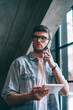 © BullRun - Pensive male user in spectacles for provide eyes protection pondering on telephone conversation indoors, concentrated hipster guy holding touch pad and calling to customer service via cellphone
