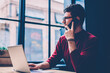 © BullRun - Serious male freelancer calling to operator for checking account balance while sitting at desktop with modern netbook, young hipster guy working with laptop device connected to cafeteria wifi