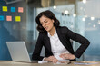 © Liubomir - Woman in office experiencing discomfort while using laptop. Signs of stress or stomach pain. Formal business attire suggests professional setting. Expression indicates need for relief.