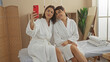 © Krakenimages.com - Two women in white robes taking a selfie in a spa center, showing a friendly moment in a wellness indoor setting.