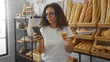 © Krakenimages.com - Woman shopping in a bakery while using a smartphone and holding a croissant with various breads on shelves in the background