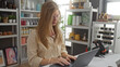 © Krakenimages.com - Young woman working on a laptop in a home decor store with blonde hair and glasses, surrounded by various decorative items and indoor plants on shelves