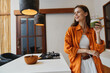 © SHOTPRIME STUDIO - Young woman in an orange shirt holding a green smoothie in a bright kitchen, showcasing healthy lifestyle, modern design, and cheerful emotions