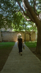 Wall Mural - Woman walking under trees on a stone path in a sunny park in palma, mallorca, spain, wearing a hat and holding a handbag near old stone walls and an arched gateway