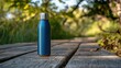 © Дмитро Жалдаков - A sleek blue stainless steel water bottle stands alone on a wooden walkway surrounded by lush greenery and dappled sunlight.