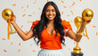 © Evgenii Starkov - A joyful female football player in an orange uniform proudly holding two golden trophies amidst confetti.