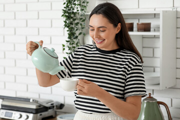 Beautiful young happy woman pouring hot water from teapot into cup in kitchen