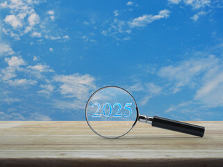 Wall Mural - Magnifying glass with 2025 text and financial graph on wooden table over blue sky with white clouds, Business happy new year 2025 research concept