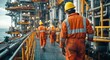 © yong - A group of workers in an oil and gas facility wearing orange overalls, yellow hard hats, and white safety boots are walking on the floor with equipment around them.