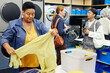 © Seventyfour - Medium shot of senior African American woman folding shirt made of delicate fabric at table checking results of washing in public laundry room with people chatting in background, copy space
