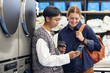 © Seventyfour - Medium shot of young Asian man showing video on phone to cheerful female friend while washing clothes and waiting for result at self service laundry