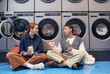 © Seventyfour - Young woman and Asian man engaged in lively conversation sitting on floor passing time, while washing machines tumbling with clothes in background in self service laundry