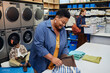 © Seventyfour - Medium shot of smiling senior African American woman packing clean clothes into bag on folding table after washing garments in public laundry room, copy space