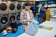 © Seventyfour - Medium shot of young Asian man packing clean clothes into bag on folding table checking results after washing garments in public laundry room, copy space