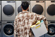 © Seventyfour - Rear view of young man dressed in plaid shirt holding basket of clothes ready for washing in tumbling machines in background in self service laundry, copy space