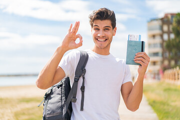 Wall Mural - Young caucasian man holding a passport at outdoors showing ok sign with fingers