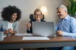 © Nenad - Three busy happy middle aged professional business man and two women executive leaders team using laptop working on computer at work desk having conversation on financial project at meeting in office.