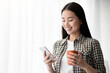 © Prostock-studio - Beautiful asian young woman with coffee mug using mobile phone, standing next to window, panorama with copy space. Cheerful lady drinking tea, chatting with boyfriend on smartphone and smiling
