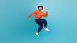 © Prostock-studio - A young African American man with curly hair is jumping joyfully against a vibrant blue background. He is wearing a casual orange sweater and blue jeans, exuding youthful energy and enthusiasm.