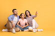 © Prostock-studio - A loving family of three shares a joyful moment against a bright yellow background. The parents sit closely with their daughter, who points at copy space while they all smile