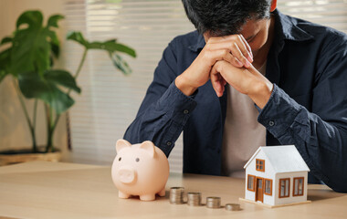 stressed man holding his forehead, surrounded by a piggy bank, coin stacks, and a house model. captu