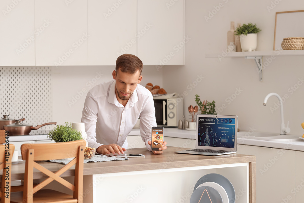 Young man using mobile phone and laptop with smart home application at table in kitchen