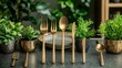 © Orata - Gold flatware is displayed upright on a dark tabletop. Small green plants in gold pots are interspersed between the utensils. A shallow plate sits behind the arrangement.